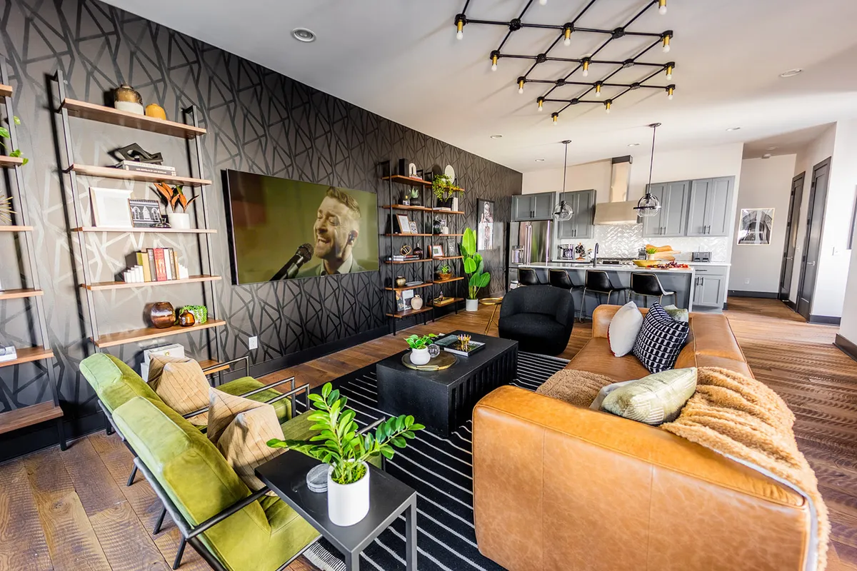 An expansive open-concept living/dining/kitchen area, showcasing a large sectional, green accent chairs, shelves with decor and books, a television displaying a performer, and the kitchen visible in the background. The space feels inviting, contemporary, and designed for both relaxation and entertainment.