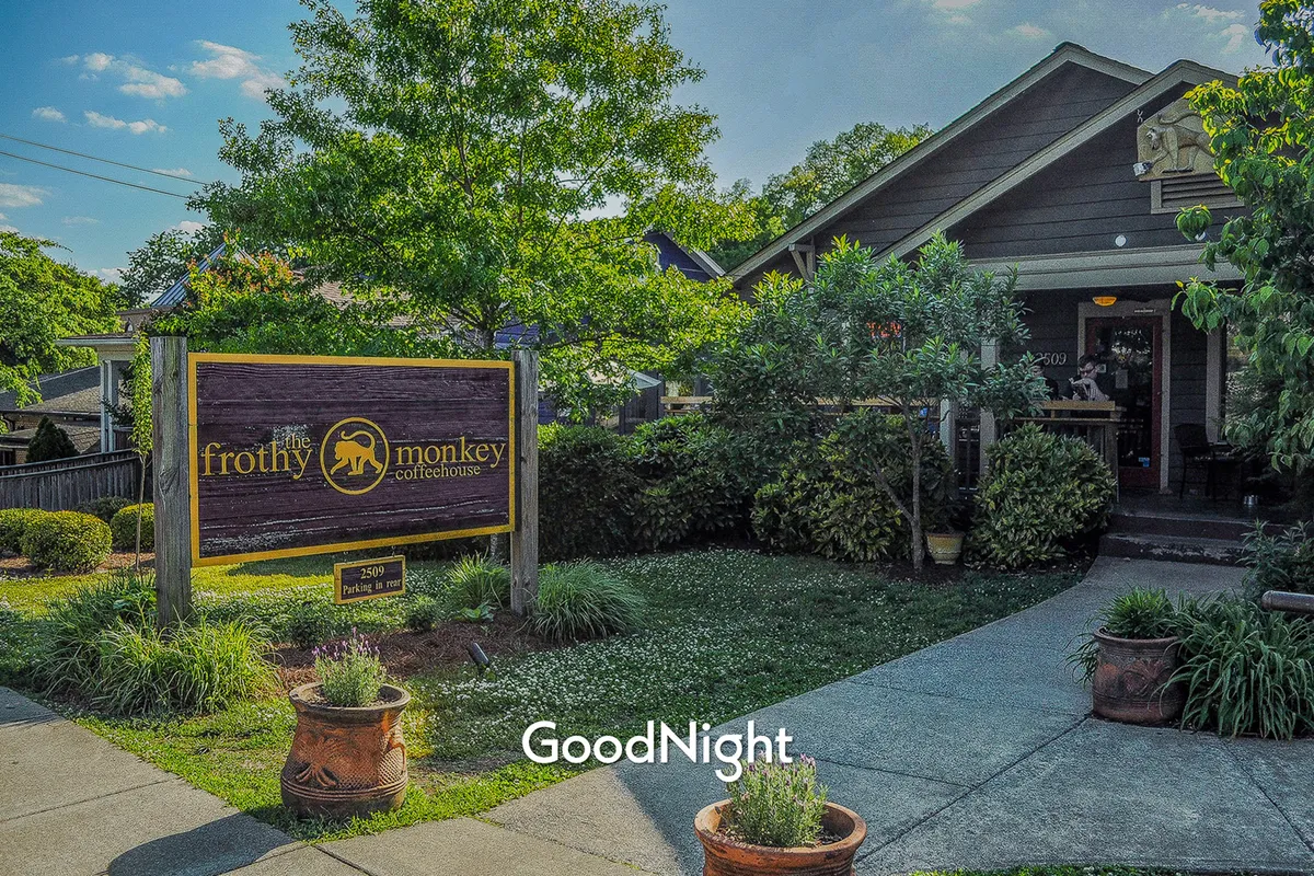 The cheerful exterior of Frothy Monkey coffeehouse, featuring its recognizable signage, leafy green foliage, and a cozy front porch perfect for lingering with friends. The setting invites relaxed conversation and community connections.