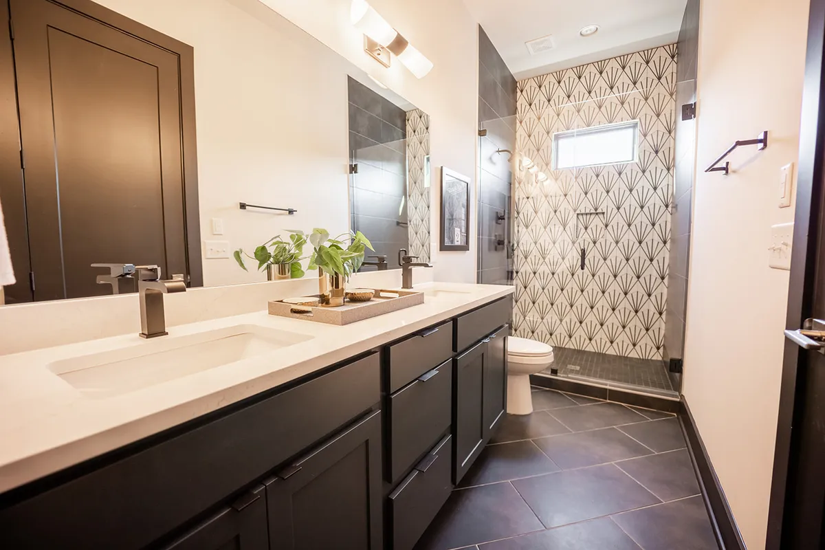 A spa-inspired en-suite bathroom with dual sinks, geometric tile shower, modern fixtures, and a potted plant. Morning light streams in, creating an inviting and rejuvenating environment for self-care and relaxation.