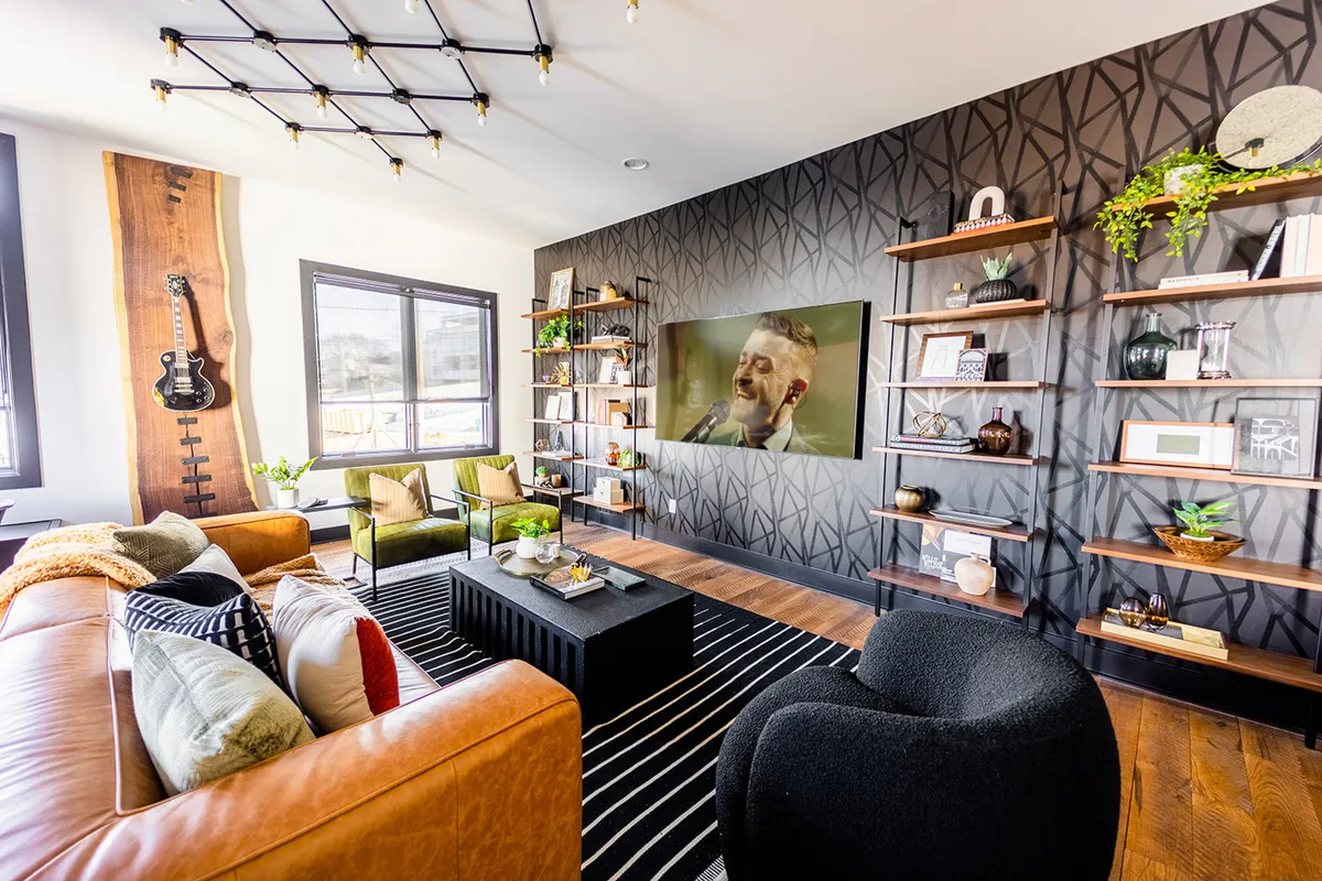 The hero image showcases a stylish, open-concept living room with a plush leather sofa, a sleek black coffee table, and curated shelves adorned with plants and decor. The centerpiece is a large TV displaying a musician, hinting at the home's musical Nashville spirit. The space feels vibrant and inviting, perfect for relaxing, entertaining friends, or soaking in the city's creative energy.