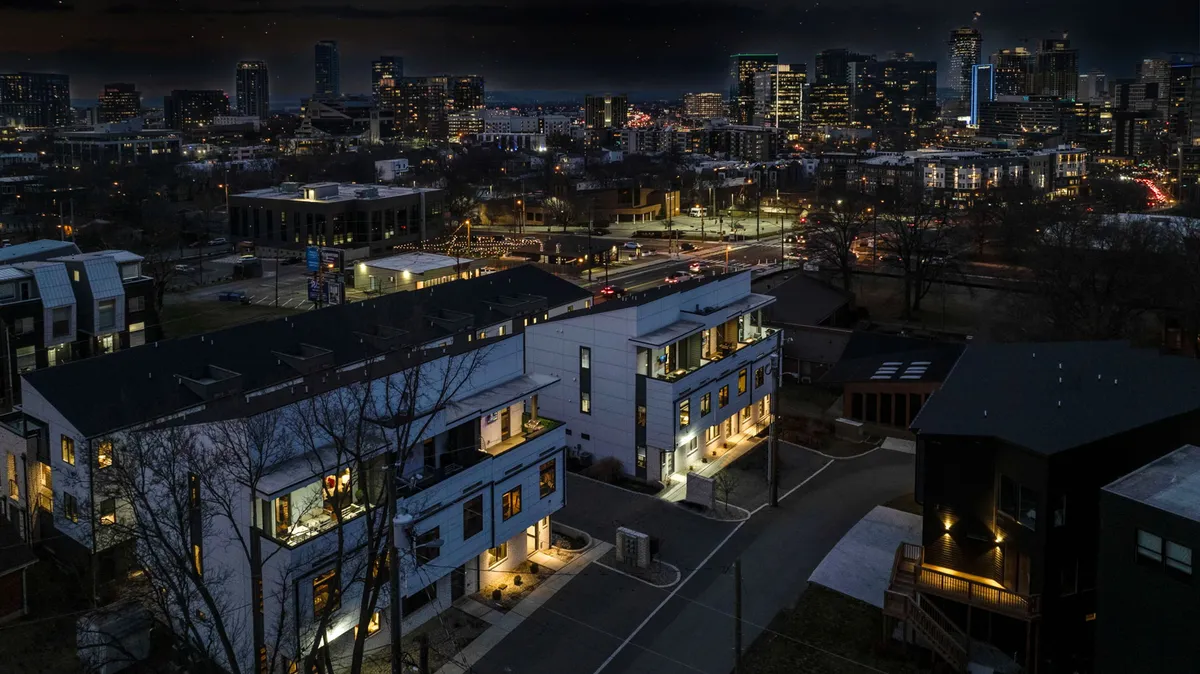 This image captures modern townhouses set against the city skyline, with street lights and a small park area visible. The urban setting reflects proximity to major business centers and the vibrancy of city life while suggesting a peaceful retreat at the end of the day.