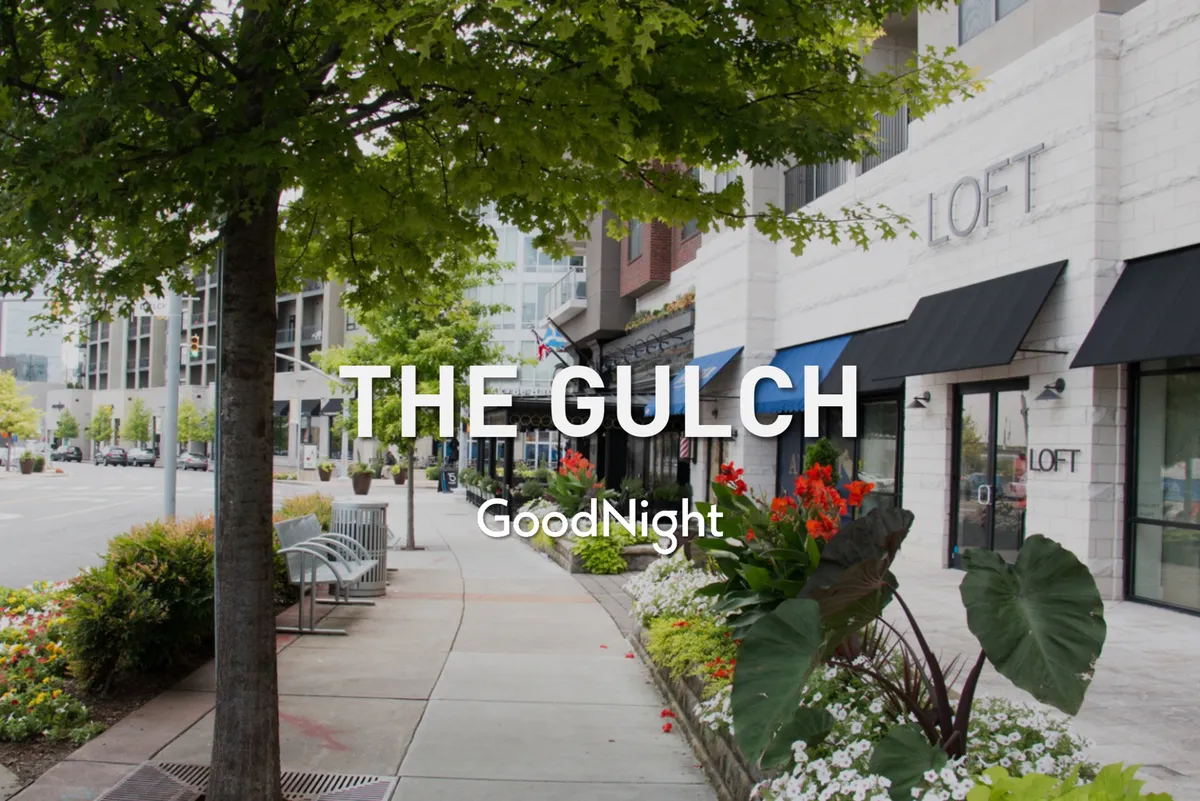 A charming streetscape featuring storefronts, flower beds, benches, and mature trees. The scene is lively yet welcoming, with people strolling and interacting in the neighborhood.