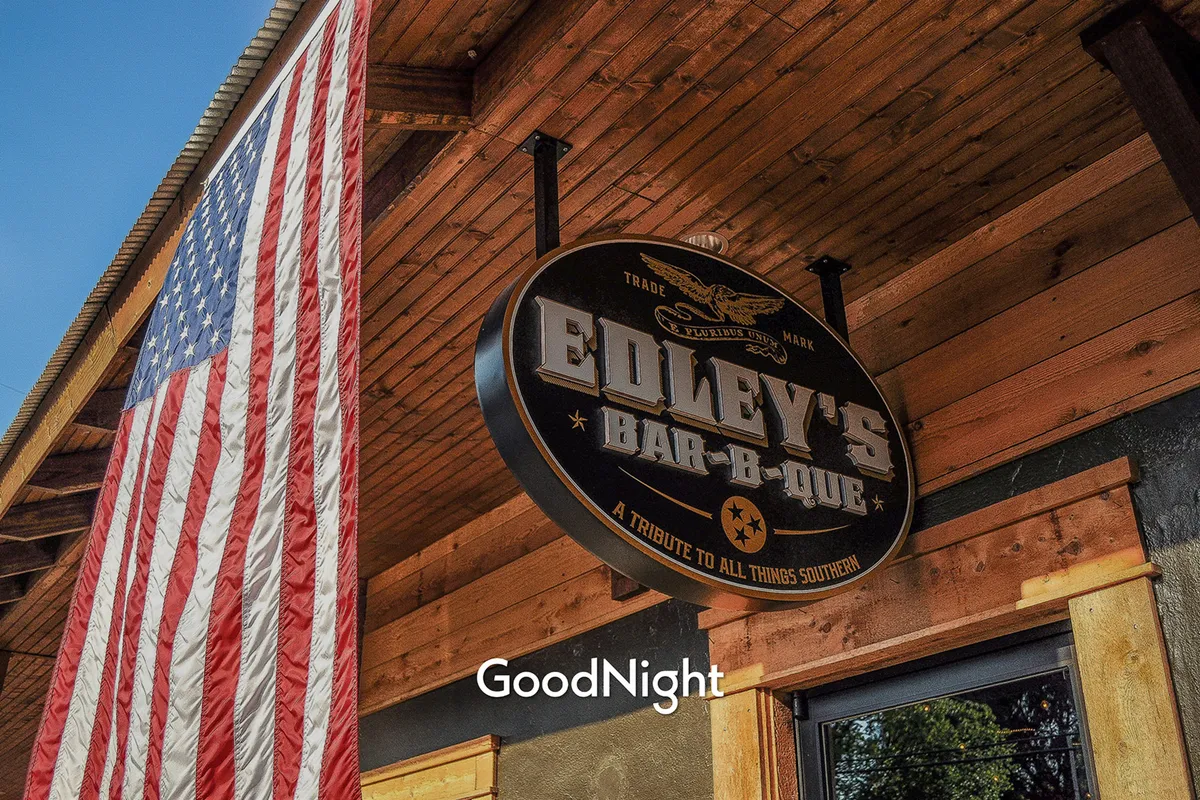 The exterior of Edley’s Bar-B-Que, marked by its welcoming signage and the American flag, signaling a beloved spot for southern cuisine. People are drawn in by the promise of classic Nashville BBQ and the lively, communal atmosphere.