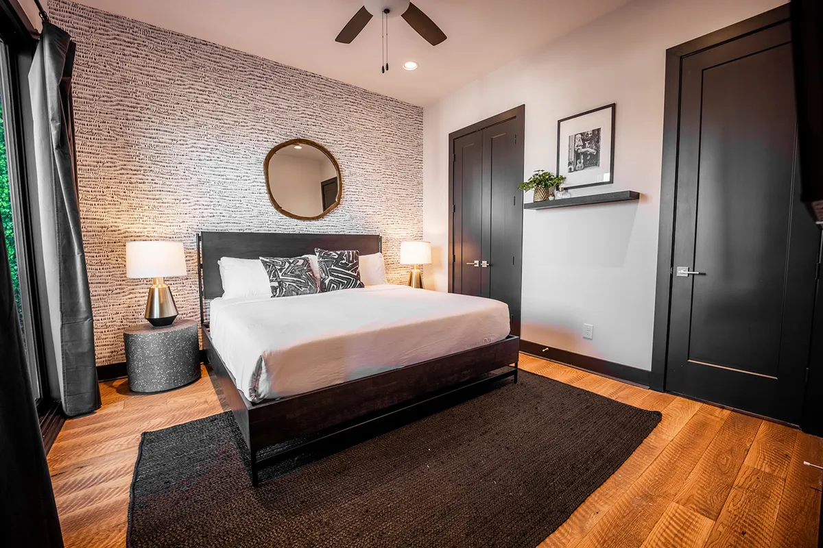 A stylish bedroom featuring a plush bed with decorative pillows, matching nightstands, sleek decorative mirrors above, and a textured accent wall. The room is bathed in natural light, creating a tranquil and inviting ambiance.