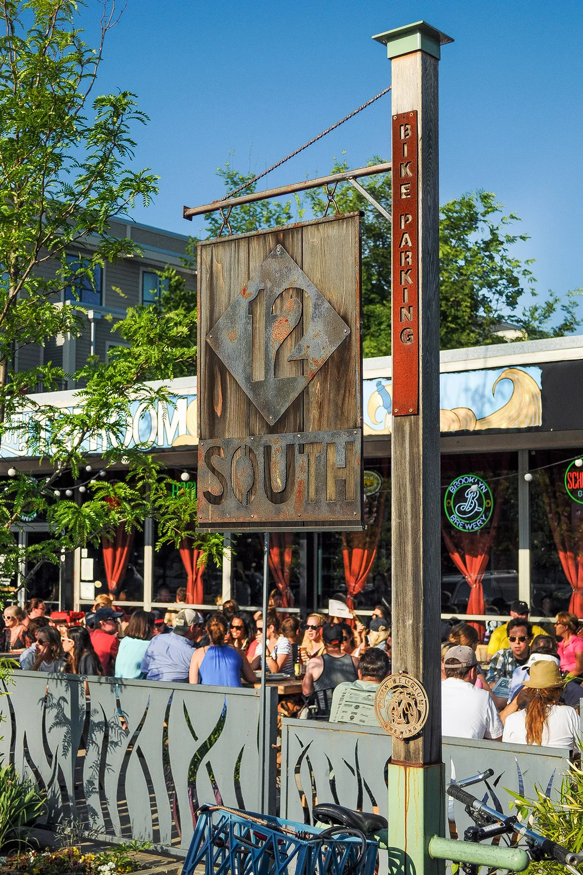The photo showcases a lively street in the 12 South neighborhood, with the recognizable '12 SOUTH' signage, a bustling crowd, and outdoor dining spaces. Decorative fencing and sunlit facades evoke a sense of community and vibrant local culture.