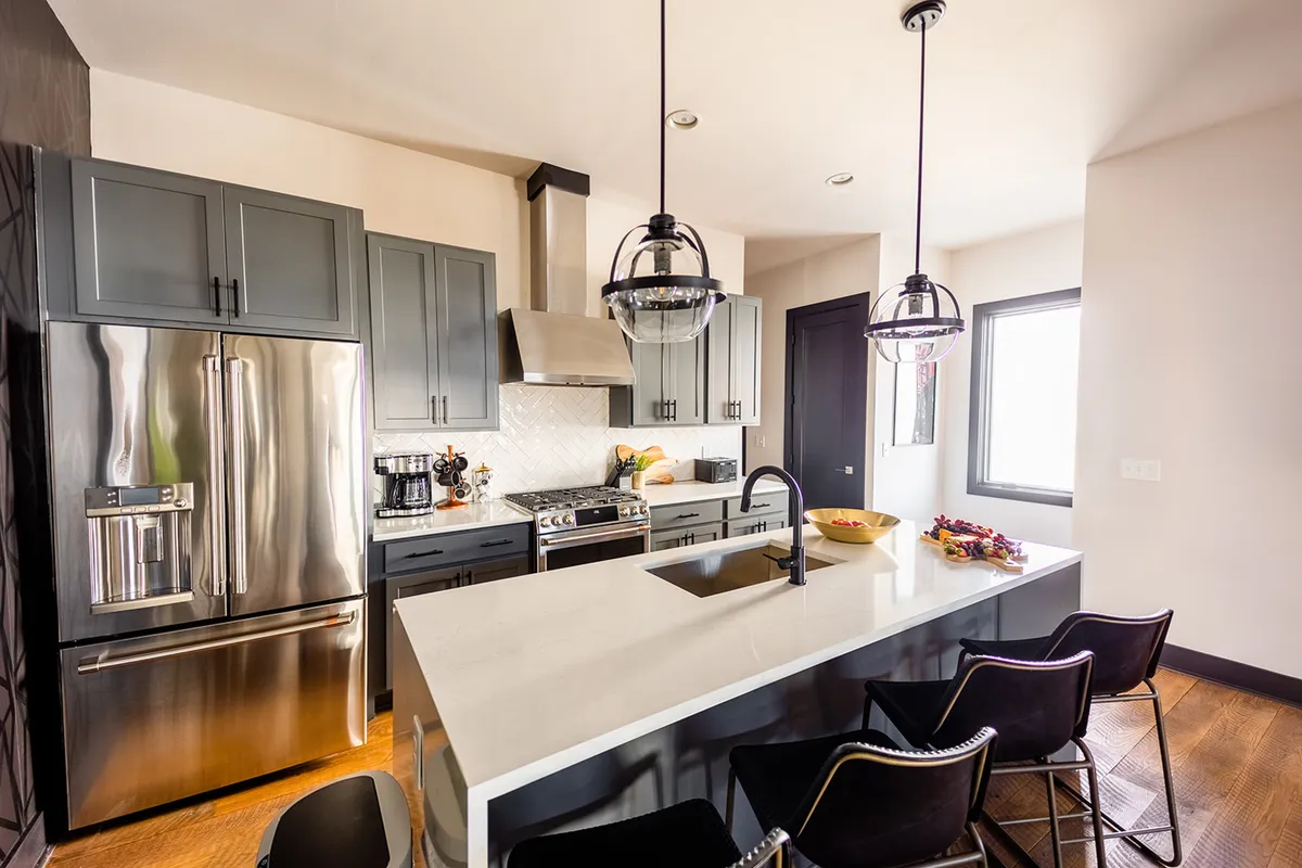 This image features a kitchen with a central island countertop, professional stainless steel appliances, and modern lighting fixtures. The overall space is open and contemporary, suggesting both functionality and style. The scene radiates warmth and welcomes both cooking and entertaining, with plenty of space for social interaction.