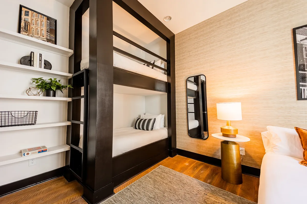 A playful and inviting bedroom featuring a sturdy bunk bed structure, a stylish mirror, and a table lamp. The design and layout are well-suited for children or groups, providing both comfort and a sense of fun.
