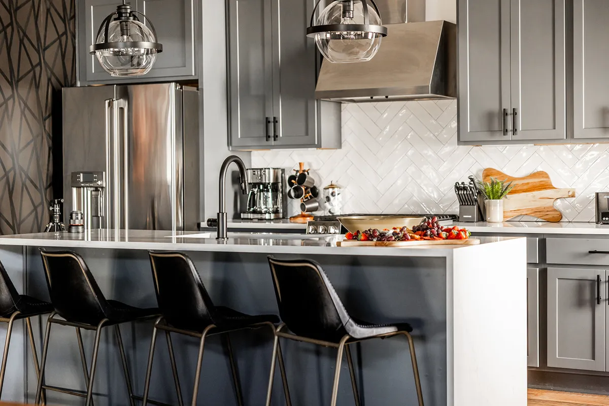 A bright, welcoming kitchen scene featuring an island countertop with fresh fruit, stylish pendant lights, textured backsplash, and modern stainless appliances. The setup is perfect for gathering, preparing meals, or enjoying casual conversations.